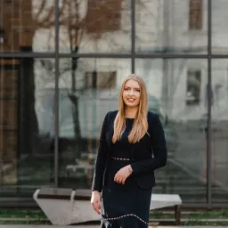 Profesjonalnie ubrana blond kobieta uśmiecha się na tle nowoczesnego budynku z przeszkloną fasadą.