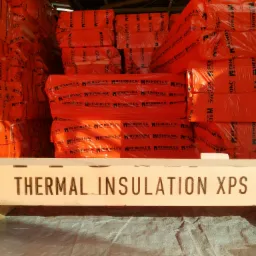 Pomarańczowe paczki materiału izolacyjnego PENOPLEX, ułożone w stosy w magazynie. Na pierwszym planie biała belka z czarnym napisem: THERMAL INSULATION XPS.