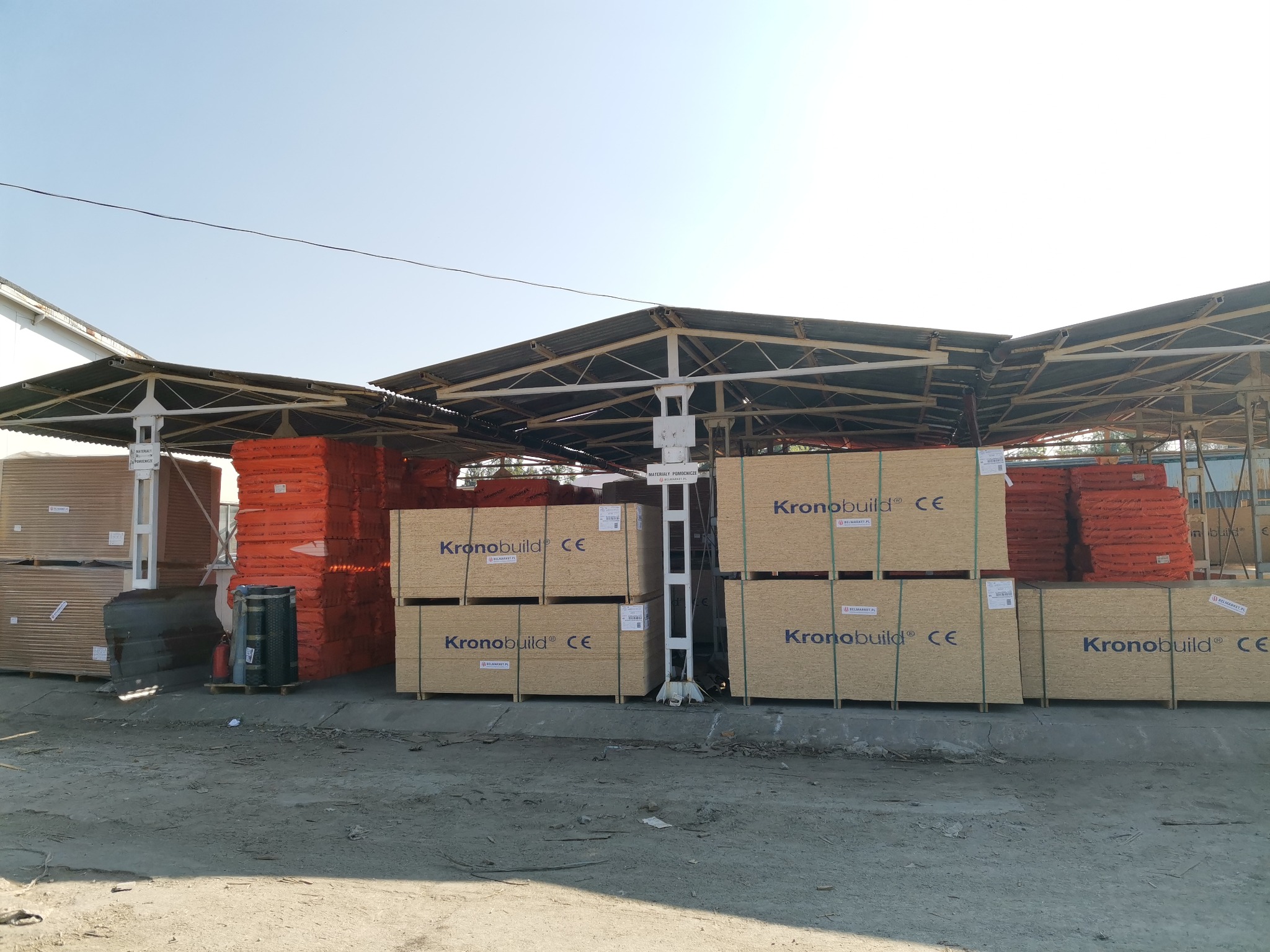 Składowane płyty OSB marki Kronobuild, widoczne palety z pomarańczową tarcicą budowlaną, całość pod zadaszeniem na placu składu budowlanego.