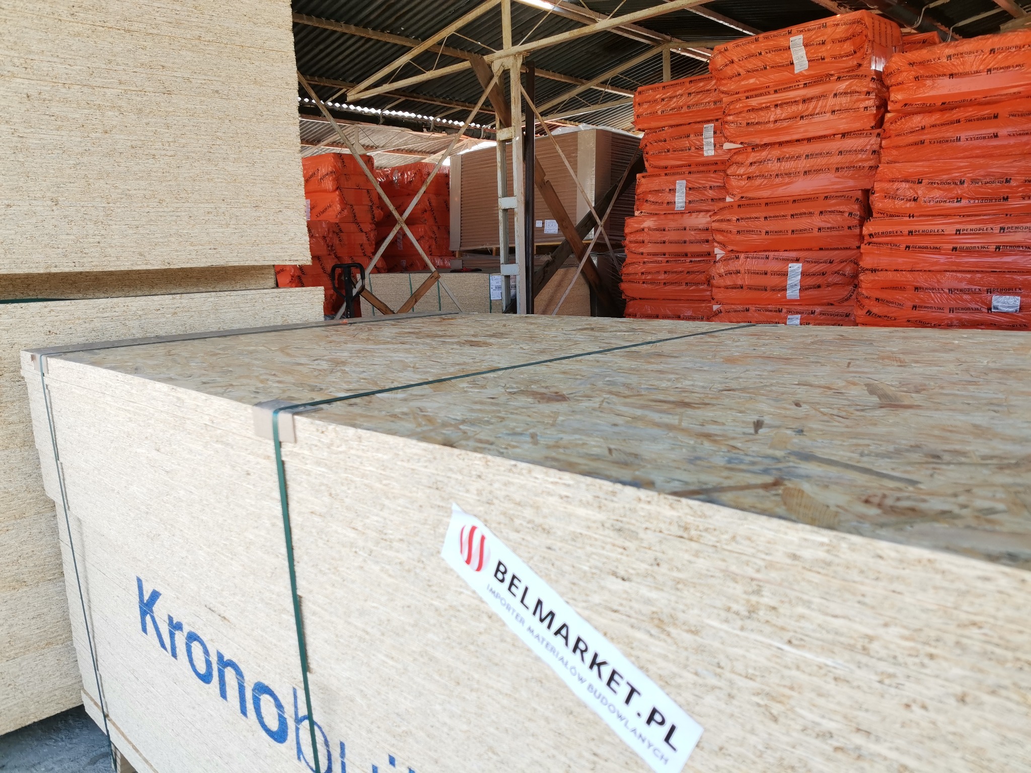 Składowane płyty OSB marki Kronobuilding z niebieskim nadrukiem, zabezpieczone zielonymi taśmami, na wierzchu naklejka Belmarket.pl, w tle widoczne pomarańczowe paczki materiałów izolacyjnych.