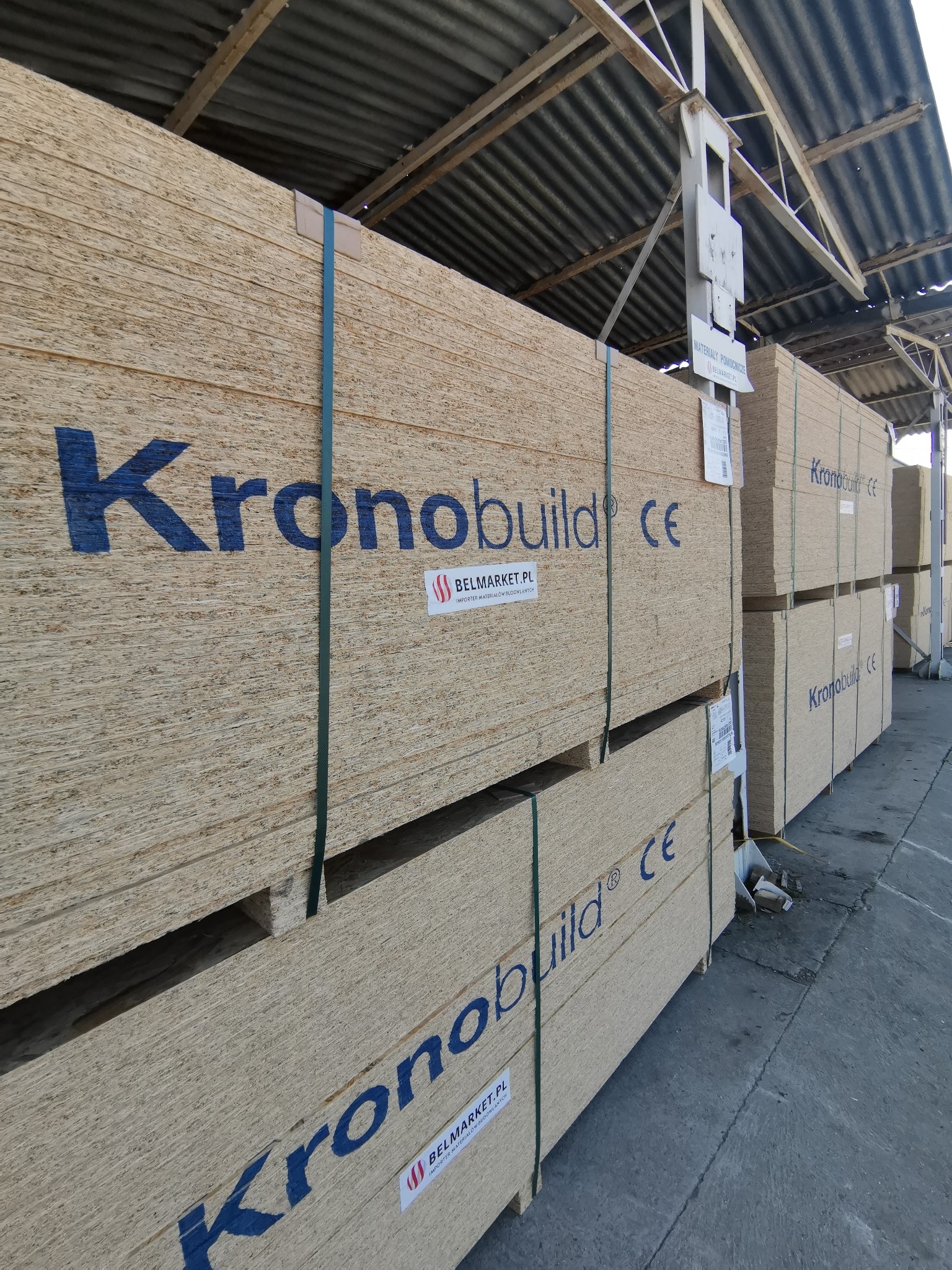 Sztaple płyt drewnopochodnych marki Kronobuild, spięte zieloną taśmą, składowane pod zadaszeniem magazynu.