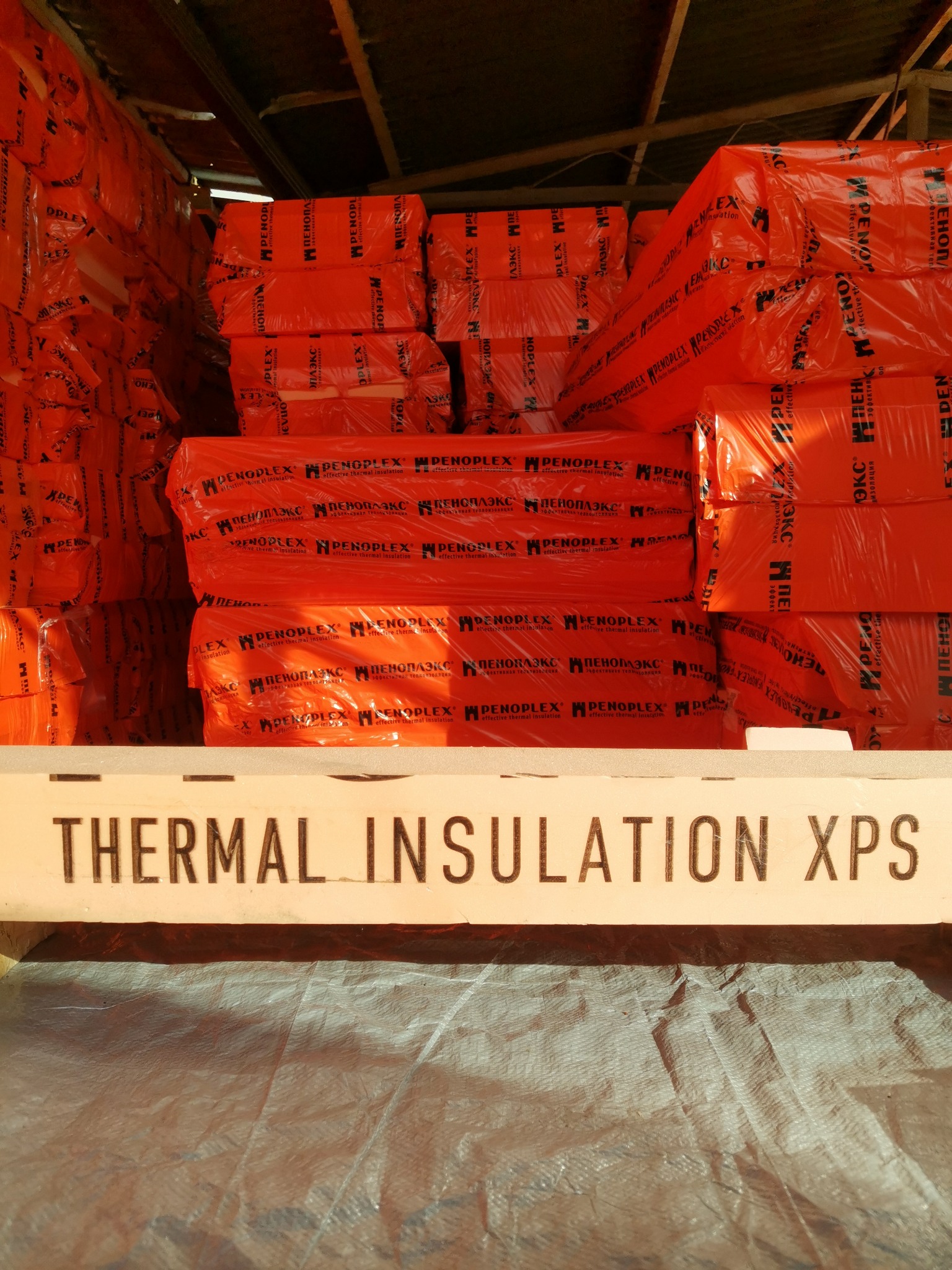 Pomarańczowe paczki materiału izolacyjnego PENOPLEX, ułożone w stosy w magazynie. Na pierwszym planie biała belka z czarnym napisem: THERMAL INSULATION XPS.