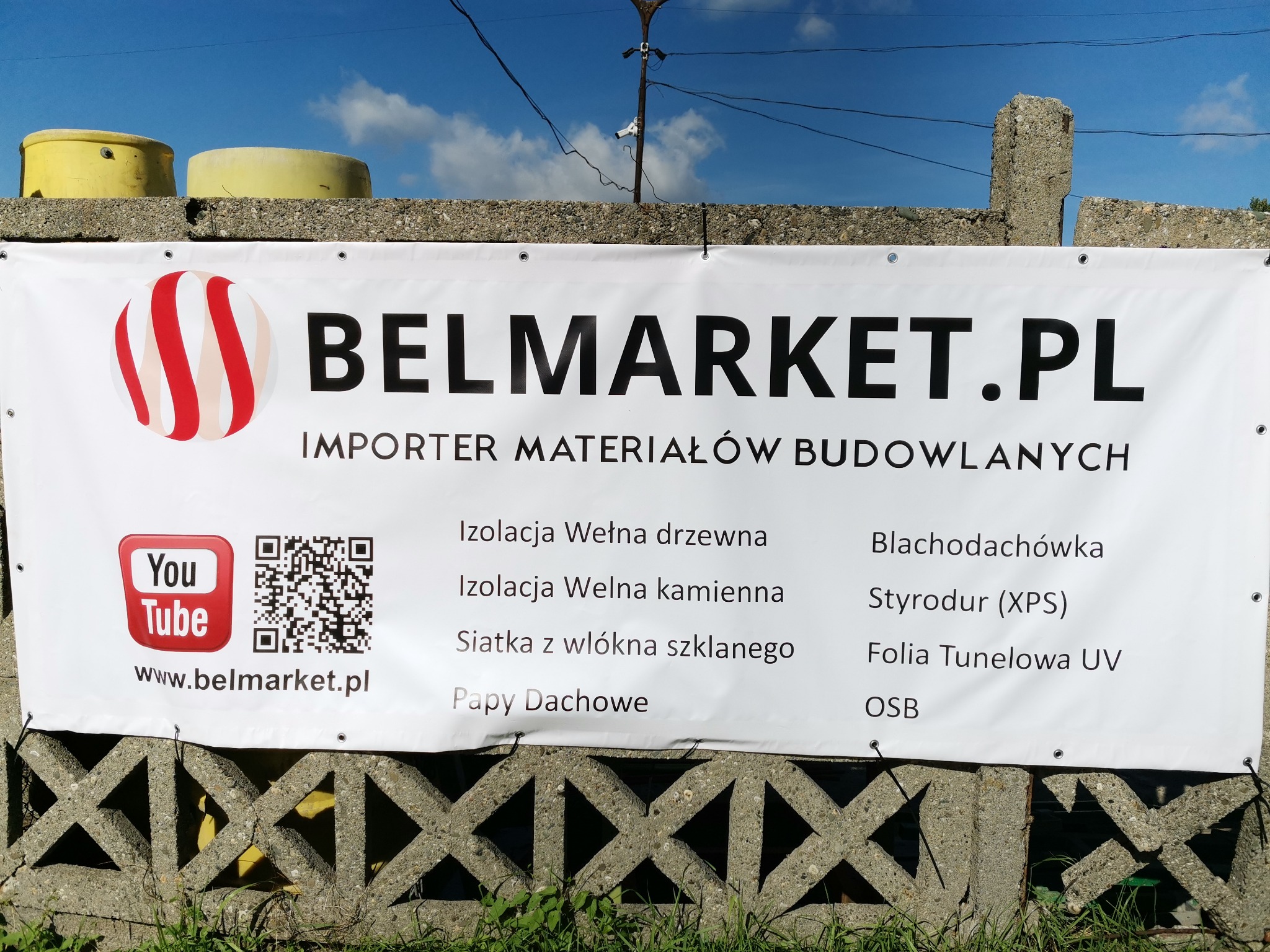 Baner reklamowy firmy Belmarket.pl, importera materiałów budowlanych, z listą oferowanych produktów, umieszczony na betonowym ogrodzeniu.