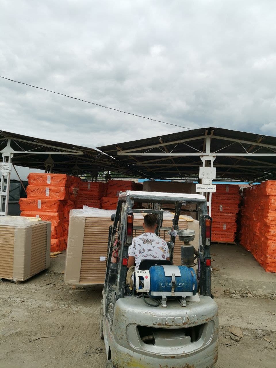 Wózek widłowy przewozi paletę z materiałami budowlanymi na terenie składu, w tle stosy pomarańczowych paczek pod zadaszeniem.