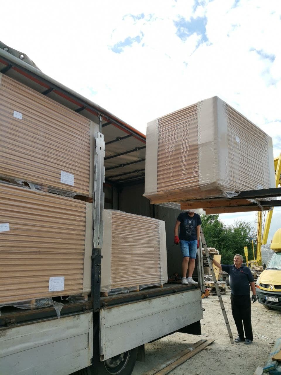 Rozładunek pakietów płyt drewnopochodnych z ciężarówki za pomocą wózka widłowego. Płyty zabezpieczone folią. Widoczni dwaj mężczyźni nadzorujący proces.