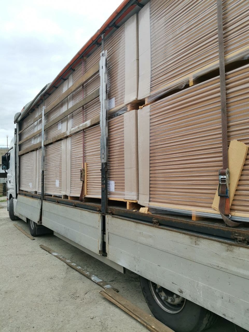 Ciężarówka załadowana paletami z blachą trapezową, zabezpieczoną folią i pasami transportowymi, widok z boku, szare niebo w tle.