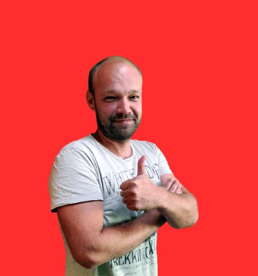 Mężczyzna z brodą w jasnej koszulce, z uniesionym kciukiem do góry na tle czerwonej ściany.