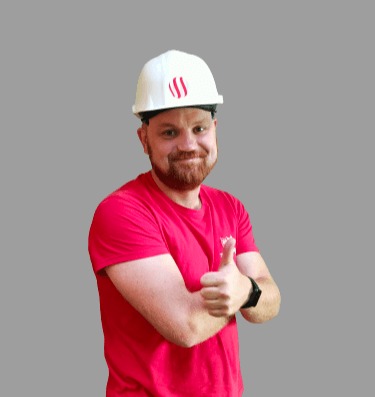 Uśmiechnięty pracownik w czerwonej koszulce i białym kasku z logo, gestykulujący kciukiem w górę.