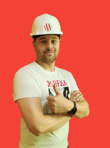 Uśmiechnięty mężczyzna w białym kasku budowlanym z logo, ubrany w białą koszulkę z nadrukiem, pokazuje kciuk w górę na jednolitym czerwonym tle.