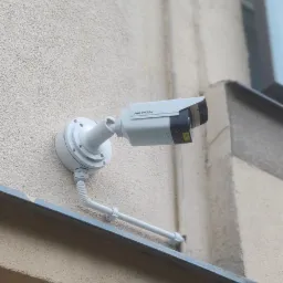 Kamera monitoringu Hikvision zamontowana na elewacji budynku, widoczne okablowanie w peszlu i rurce PCV, jasna elewacja i fragment okna w tle.
