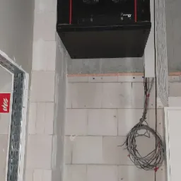 Czarna szafa rackowa zamontowana pod sufitem w surowym pomieszczeniu, widoczne kable i przewody, ściany z białych bloczków, wentylatory w obudowie, srebrna rura wentylacyjna w tle.