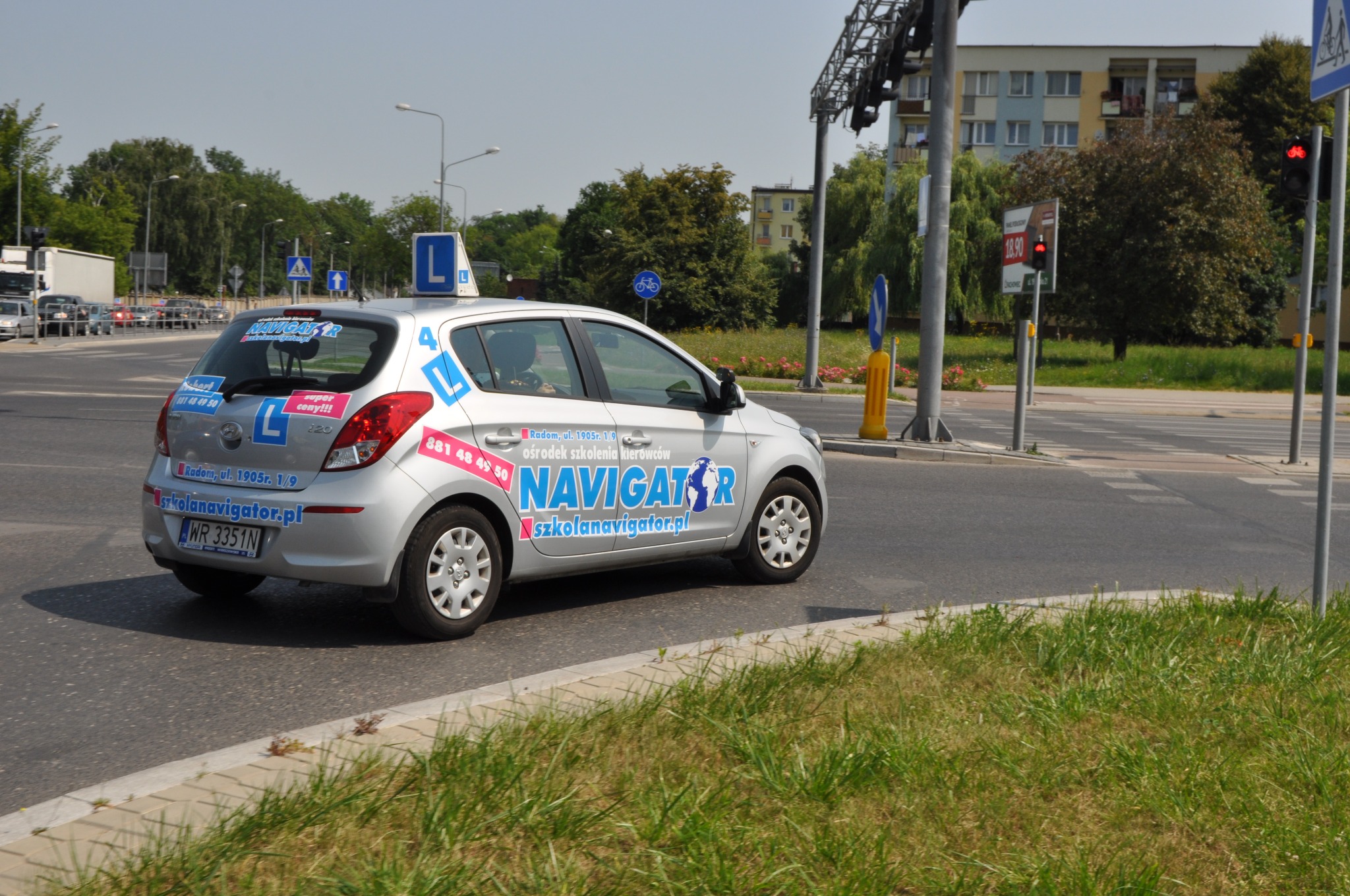 Srebrny samochód nauki jazdy z dużym 'L' na dachu i naklejkami 'NAVIGATOR' oraz numerami telefonów, skręcający na skrzyżowaniu z sygnalizacją świetlną w tle.