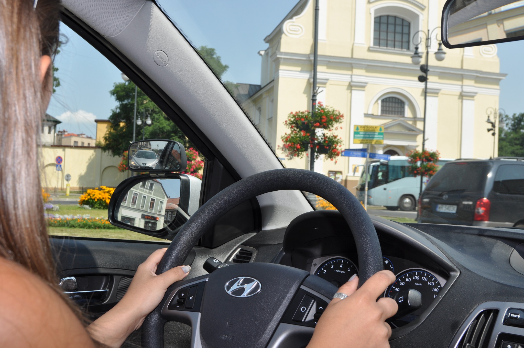 Kobieta trzyma kierownicę samochodu marki Hyundai, widok z perspektywy kierowcy na ulicę z budynkami i pojazdami, lusterko wsteczne pokazuje zbliżający się pojazd.