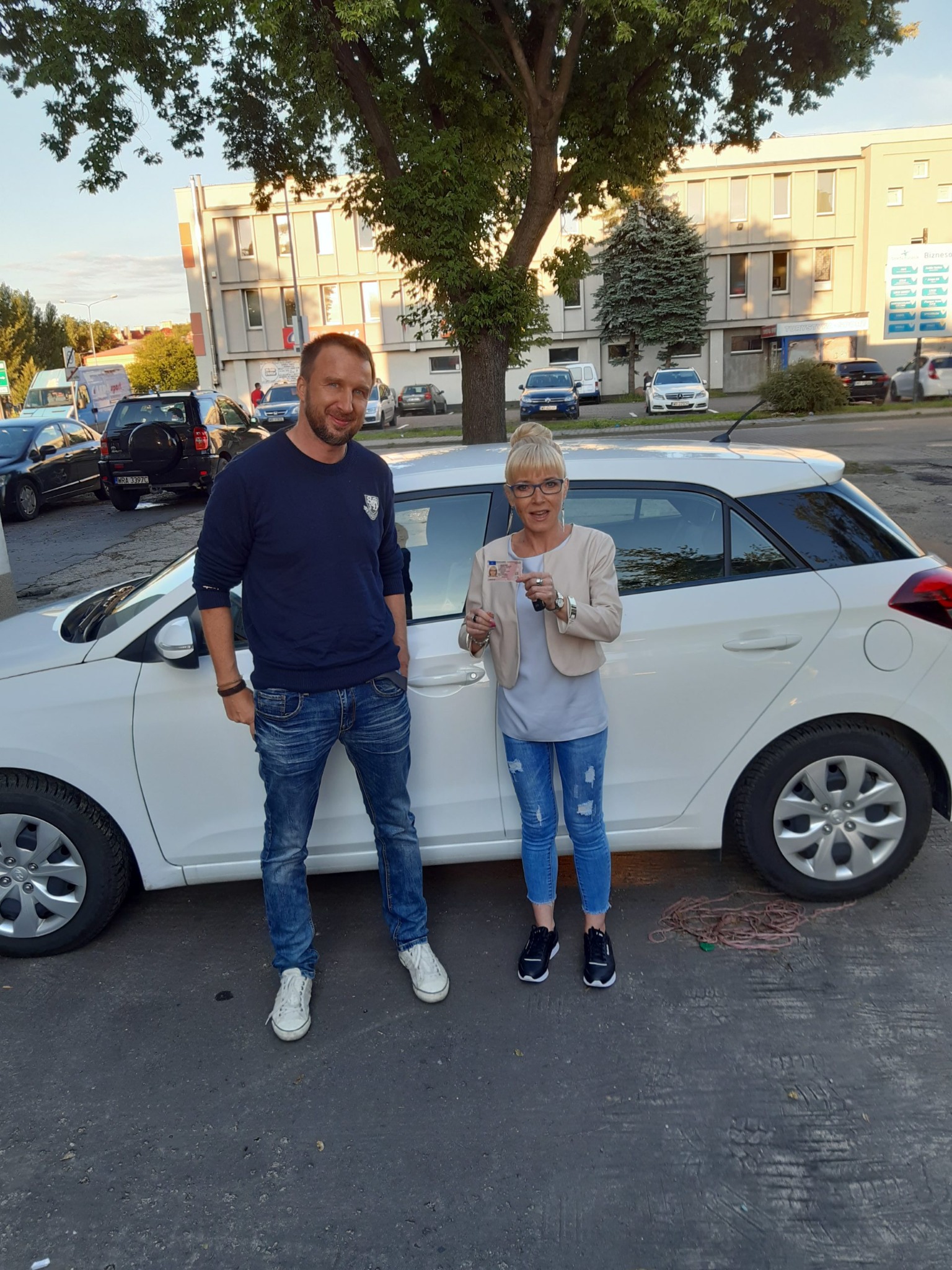 Kobieta z blond kokiem i okularami trzyma prawo jazdy, stojąc obok białego samochodu z instruktorem na tle budynku.