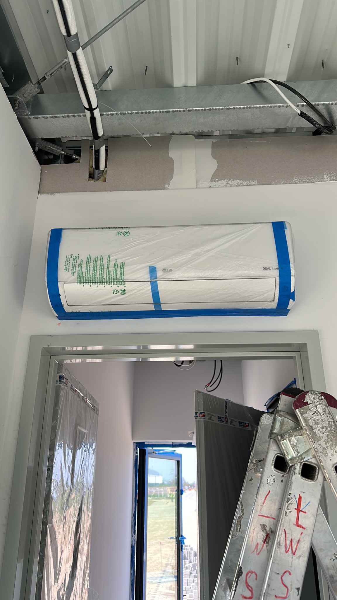 Nowo zamontowana klimatyzacja LG Dual Inverter, zabezpieczona folią i niebieską taśmą malarską, w jasnym biurze podczas remontu, widoczna drabina i elementy wykończeniowe.