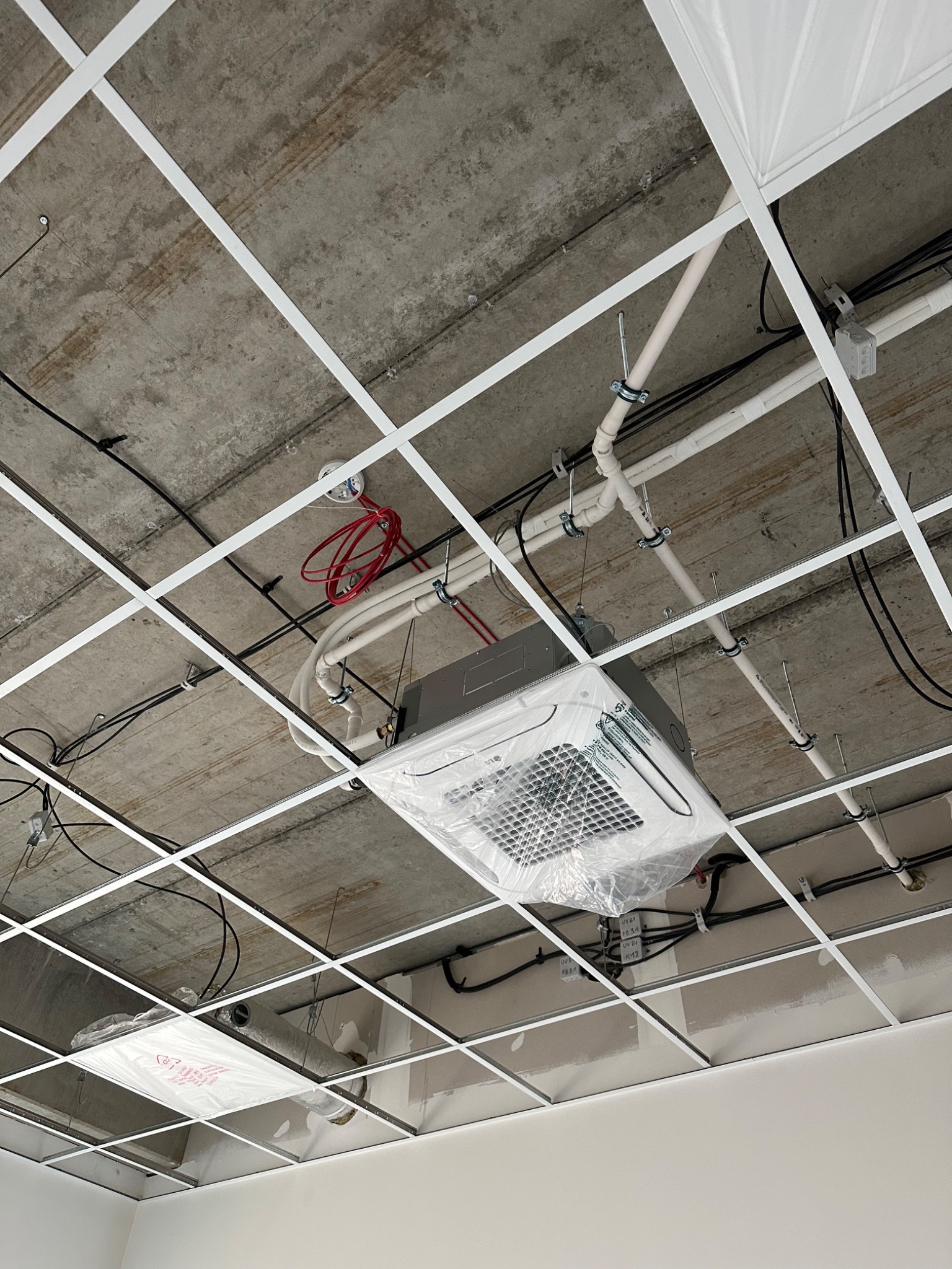 Kasetonowa jednostka klimatyzacyjna w folii ochronnej, zamontowana pod betonowym sufitem z widoczną instalacją elektryczną i rurami, częściowo ukryta za konstrukcją podwieszanego sufitu.