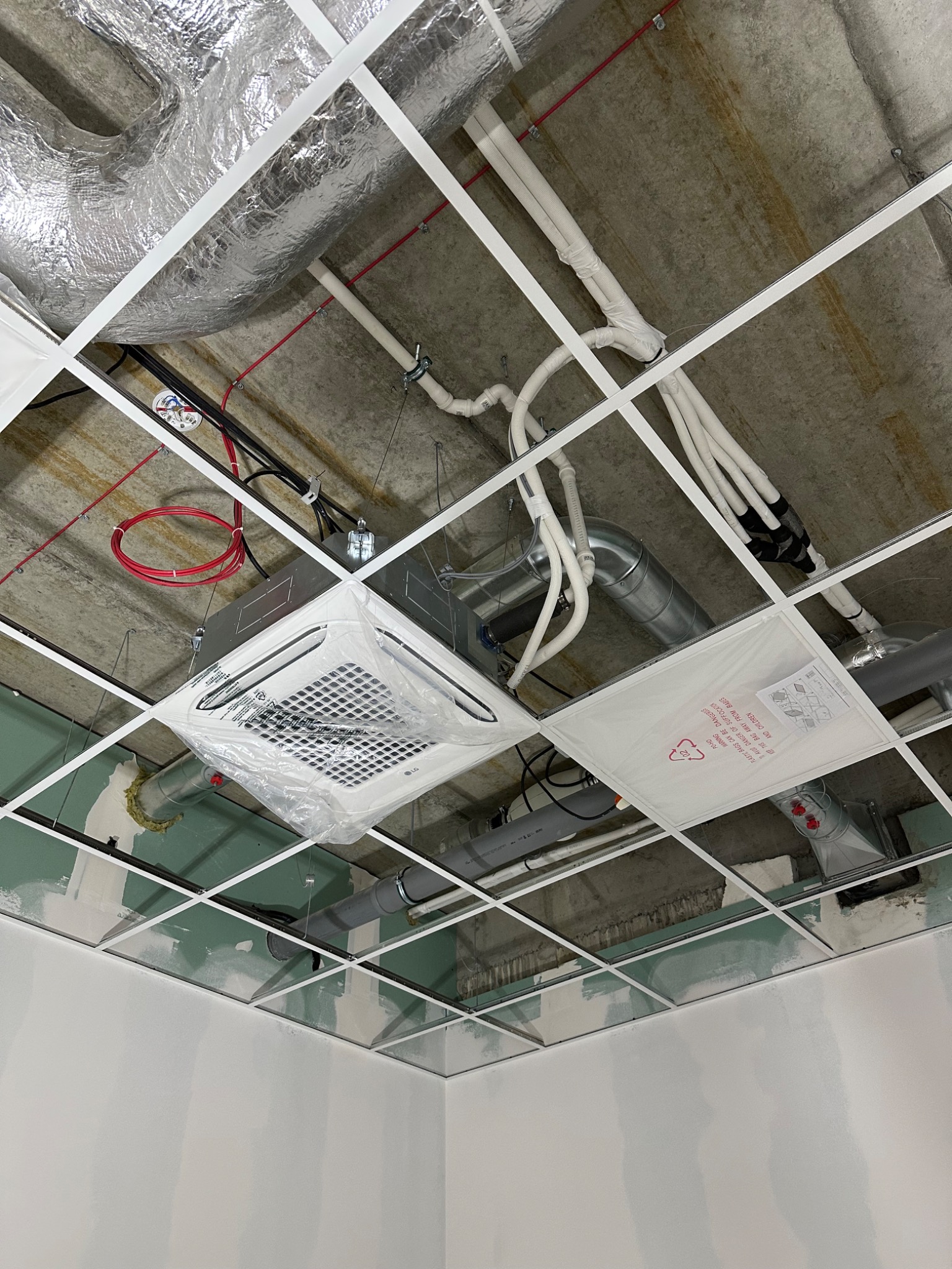 Montaż klimatyzacji kasetonowej w biurze, widoczne elementy instalacji pod sufitem podwieszanym, jednostka wewnętrzna w folii ochronnej LG, rury i kable instalacyjne, surowy stan wykończenia wnętrza.