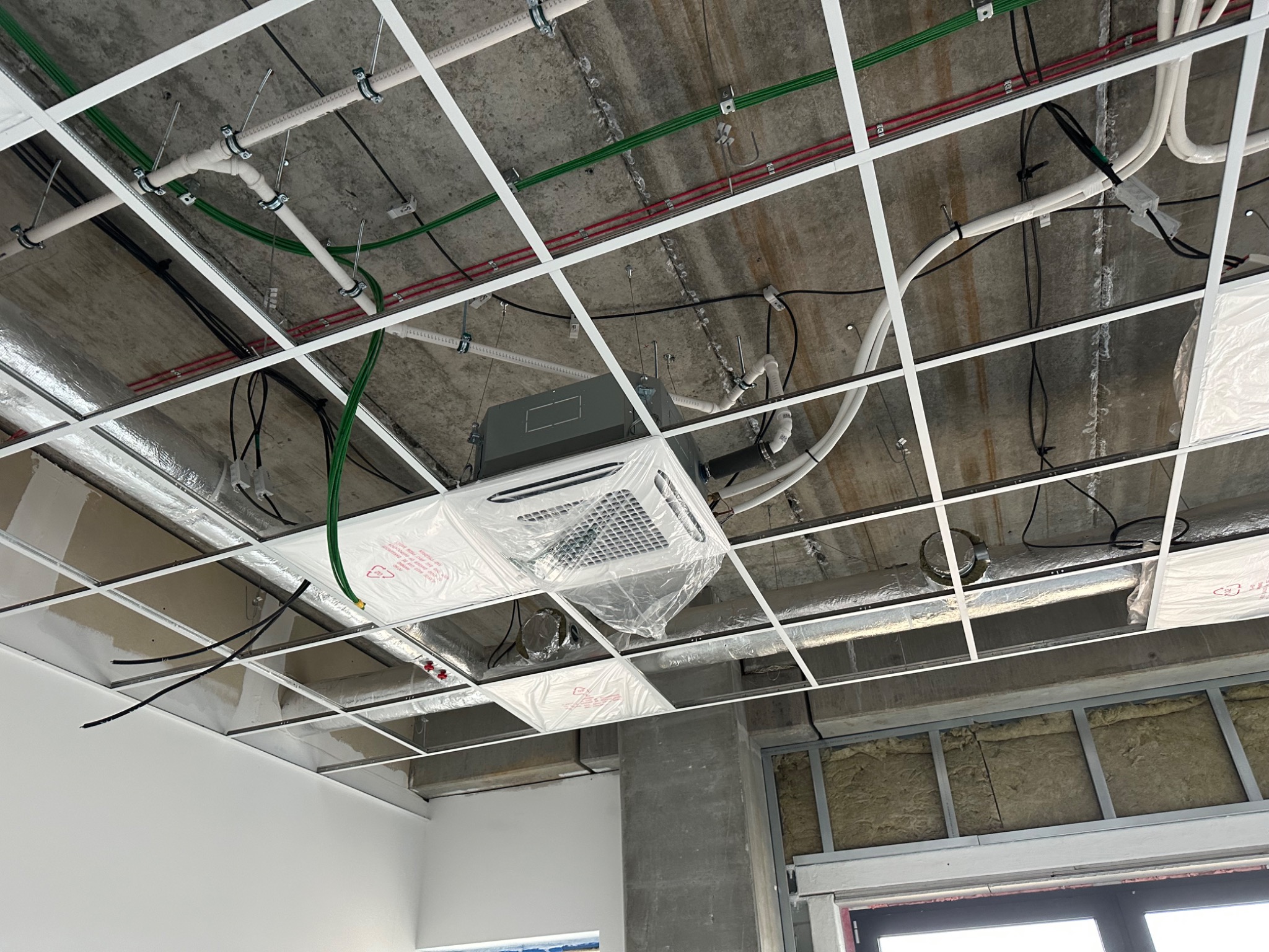 Kasetonowa klimatyzacja sufitowa w folii ochronnej, widoczna na tle surowego sufitu z instalacjami i elementami konstrukcyjnymi w trakcie montażu.