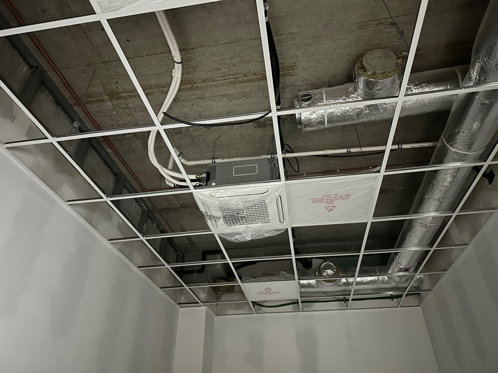 Montaż klimatyzacji kasetonowej w biurze, widoczne rury wentylacyjne i instalacja elektryczna pod sufitem podwieszanym, urządzenie jeszcze w folii ochronnej.