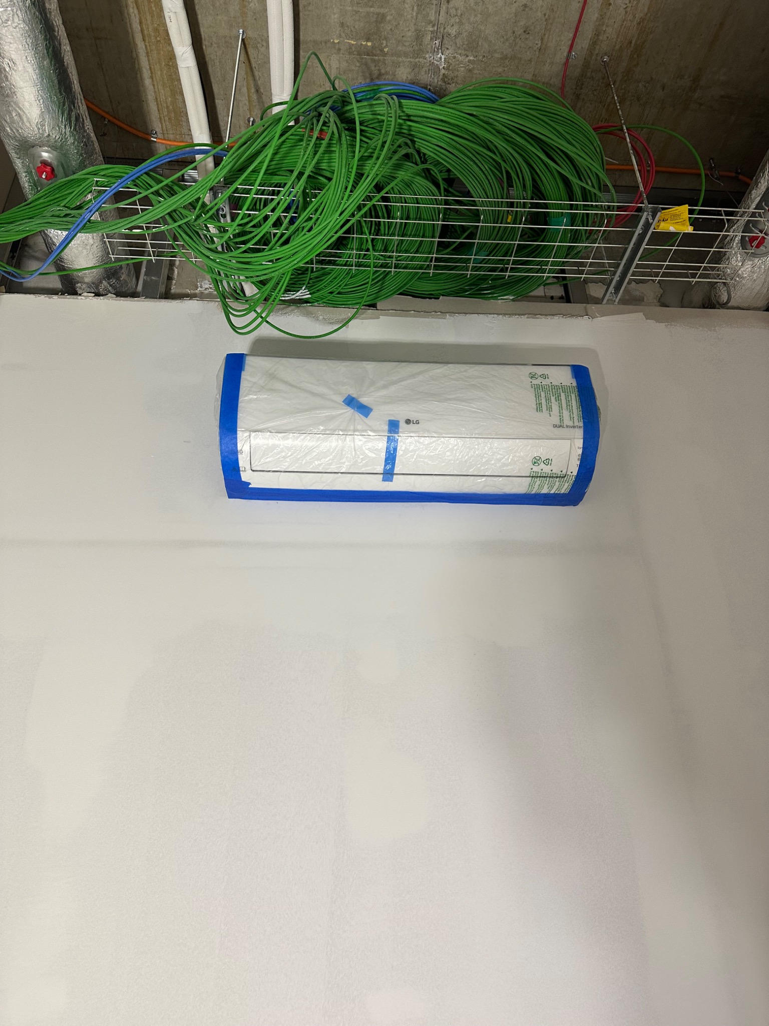 Klimatyzator LG, zabezpieczony folią i taśmą malarską, zamontowany na ścianie, z widocznymi kablami w korytku pod sufitem.