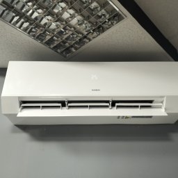 DANMAX Spółka z o.o. - Biała jednostka klimatyzacji Kaisai zamontowana na szarej ścianie pod sufitem z rastrowymi lampami.