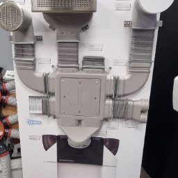 DANMAX Spółka z o.o. - Ekspozycja systemu Zehnder Comfosystems OnFloor z elementami wentylacyjnymi, w tym anemostaty i kanały, na białym panelu z czerwonym cokołem.