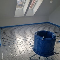 DANMAX Spółka z o.o. - Instalacja ogrzewania podłogowego w nowym domu: niebieskie rurki ułożone w pętle na srebrnej folii izolacyjnej, widok z góry na poddaszu z oknem dachowym.
