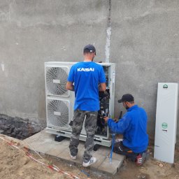 DANMAX Spółka z o.o. - Dwóch instalatorów w trakcie montażu zewnętrznej jednostki klimatyzacji Kaisai przy szarej ścianie budynku, widoczny również panel ECO HOME R32 oraz wykop zabezpieczony taśmą.