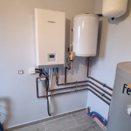 DANMAX Spółka z o.o. - Biały wiszący piec Kaisai z podłączonymi miedzianymi rurami, obok biały zasobnik wody i szary zbiornik na tle szarej ściany i podłogi z płytek imitujących drewno.