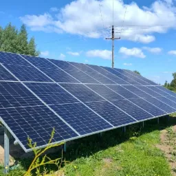 Panel słoneczny na metalowej konstrukcji ustawiony na trawie, w tle drzewa, pole i błękitne niebo z chmurami.