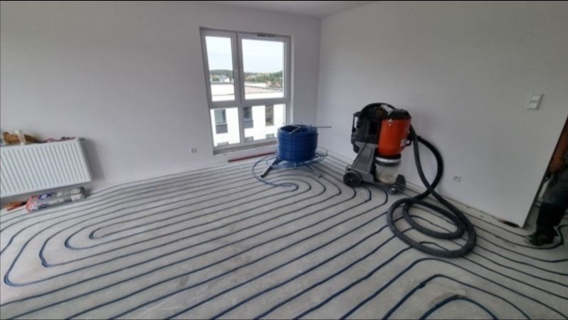 Rozłożona instalacja ogrzewania podłogowego z niebieskich rurek, widoczny rozwijak z rolką rur, odkurzacz przemysłowy i fragment grzejnika na ścianie w surowym stanie.