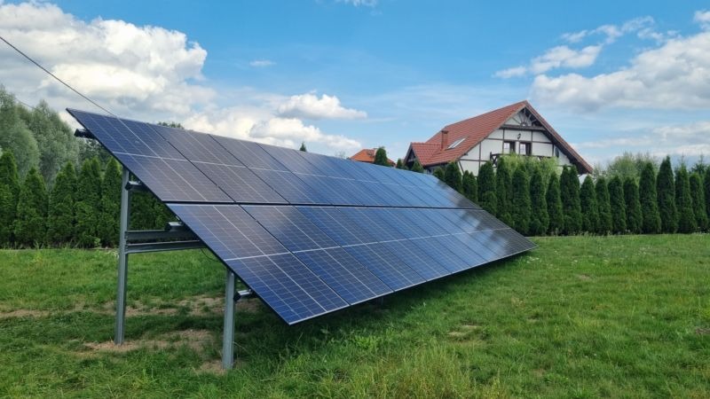 Panel słoneczny na metalowej konstrukcji w ogrodzie przed domem z czerwoną dachówką, na tle błękitnego nieba z chmurami.
