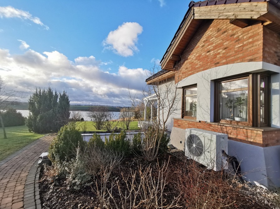 Zewnętrzna jednostka klimatyzacji Midea zamontowana przy ścianie domu z ceglaną elewacją, z widokiem na jezioro i zachmurzone niebo w tle.