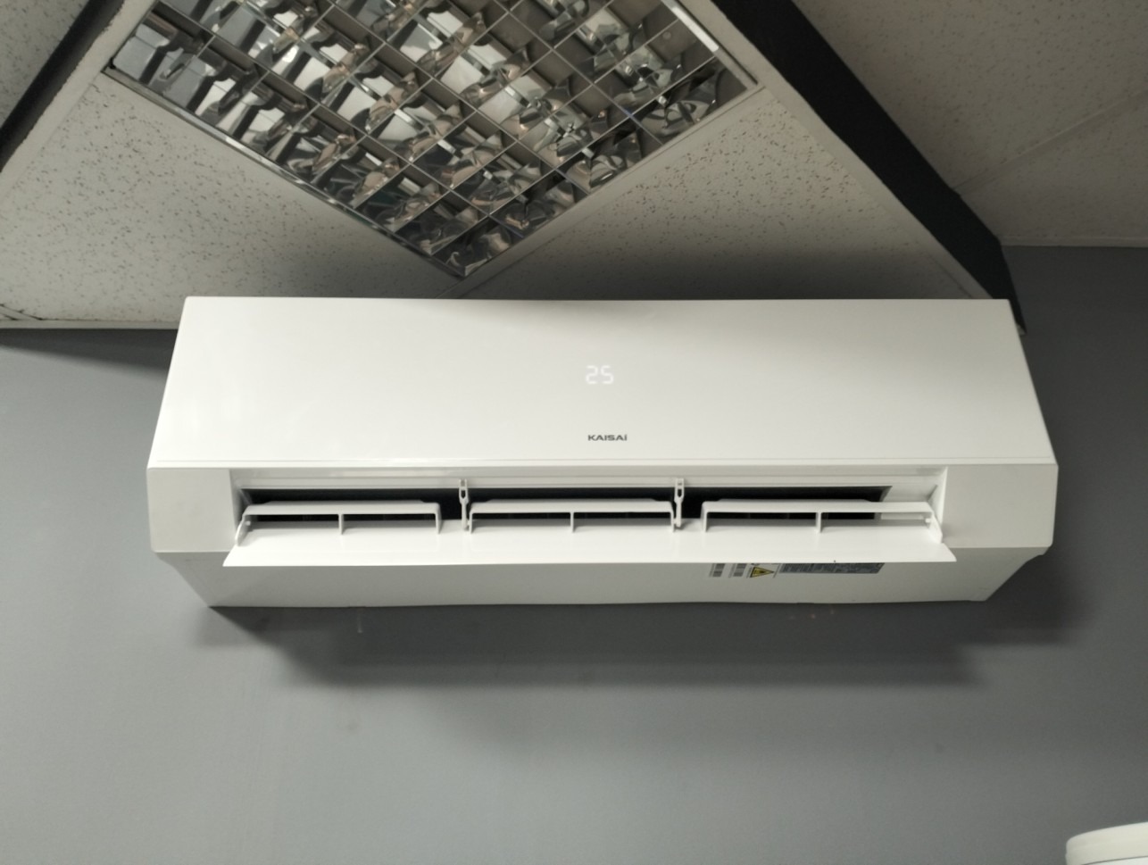 Biała jednostka klimatyzacji Kaisai zamontowana na szarej ścianie pod sufitem z rastrowymi lampami.