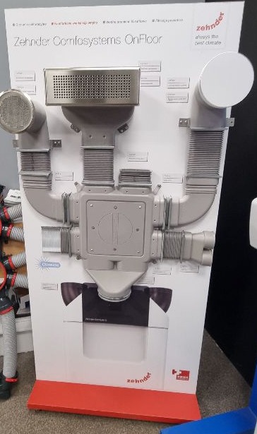 Ekspozycja systemu Zehnder Comfosystems OnFloor z elementami wentylacyjnymi, w tym anemostaty i kanały, na białym panelu z czerwonym cokołem.