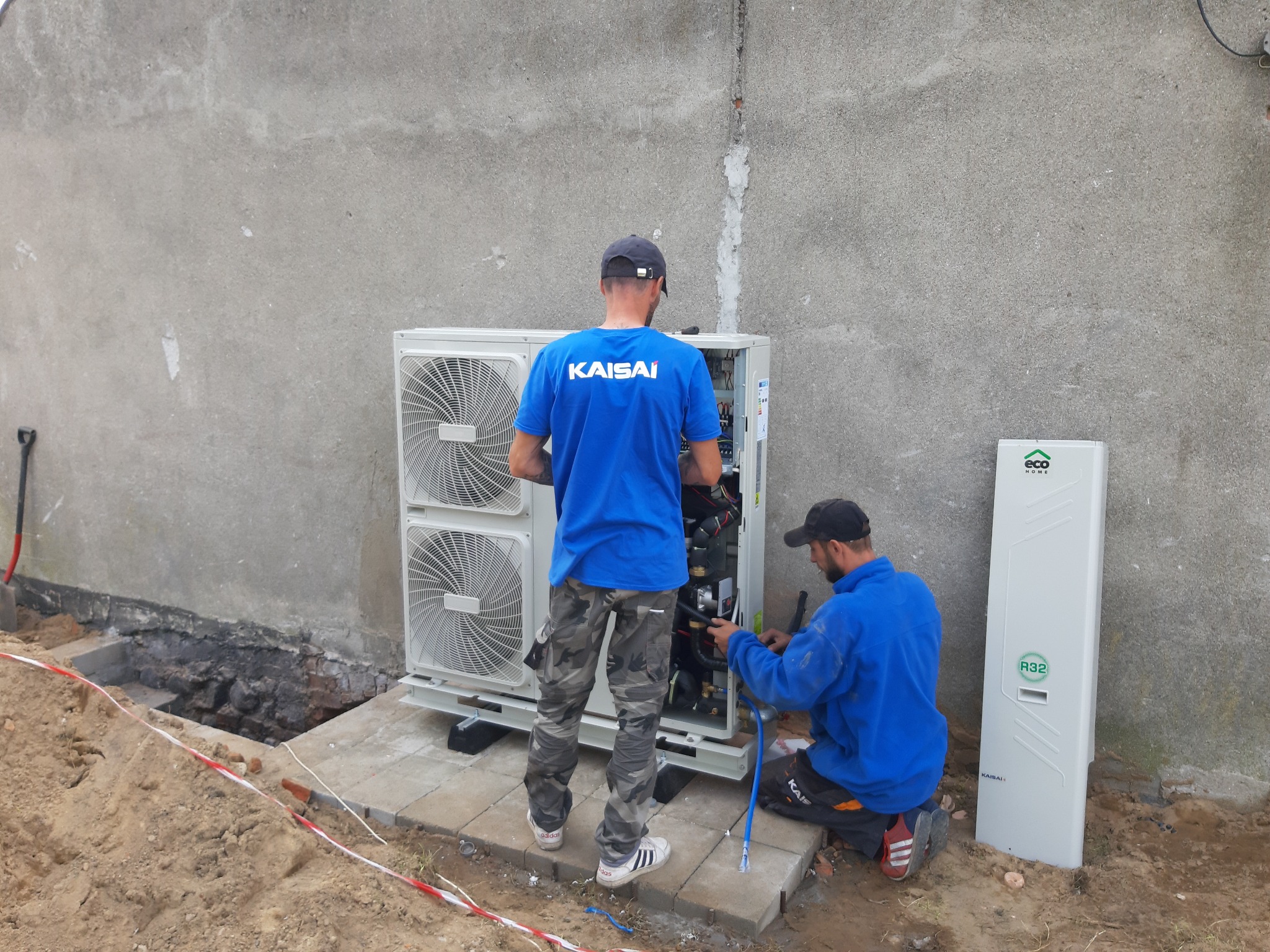 Dwóch instalatorów w trakcie montażu zewnętrznej jednostki klimatyzacji Kaisai przy szarej ścianie budynku, widoczny również panel ECO HOME R32 oraz wykop zabezpieczony taśmą.