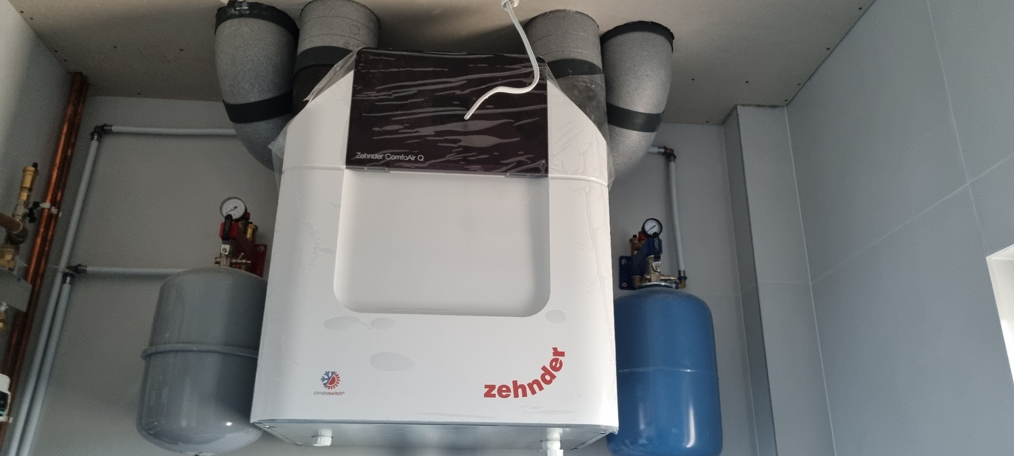 Centrala rekuperacyjna Zehnder ComfoAir Q z podłączonymi przewodami wentylacyjnymi i instalacją hydrauliczną, widoczne manometry i zbiorniki wyrównawcze.
