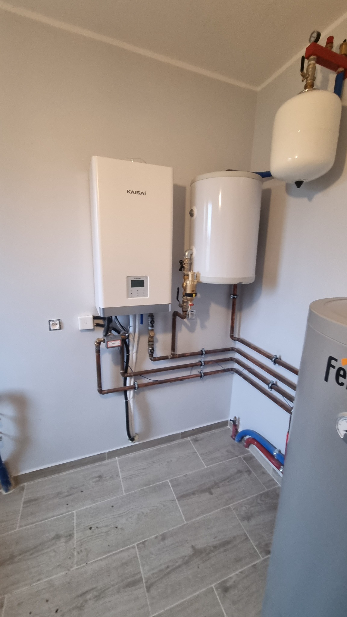 Biały wiszący piec Kaisai z podłączonymi miedzianymi rurami, obok biały zasobnik wody i szary zbiornik na tle szarej ściany i podłogi z płytek imitujących drewno.