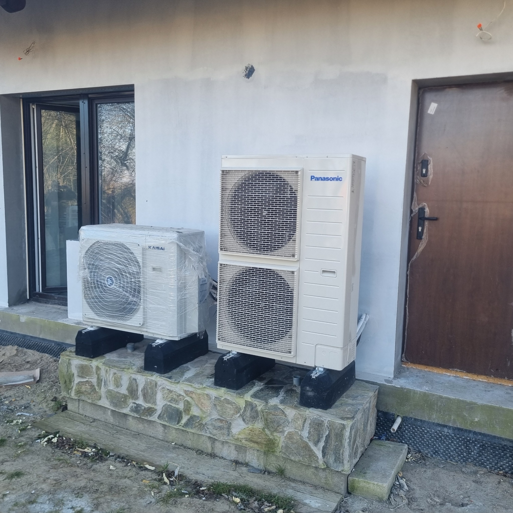 Dwie jednostki zewnętrzne klimatyzacji, marki Kaisai (w folii ochronnej) i Panasonic, zamontowane na kamiennym cokole przy budynku w trakcie wykończenia elewacji.