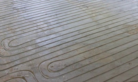 Szare rury ogrzewania podłogowego ułożone w betonowej wylewce, widok z góry pod kątem.