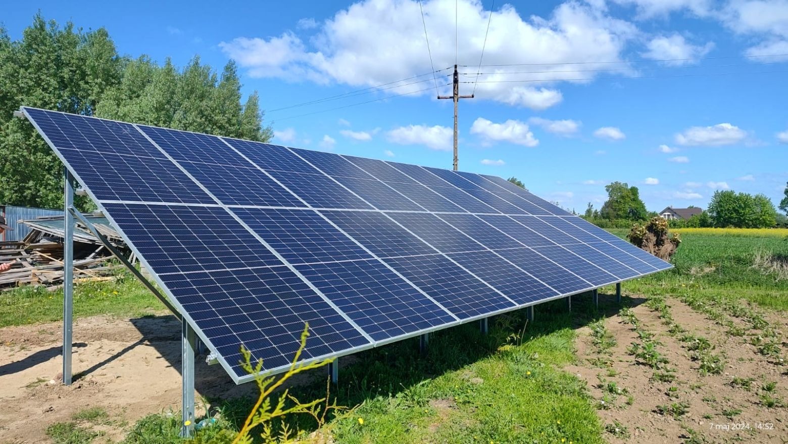 Panel słoneczny na metalowej konstrukcji ustawiony na trawie, w tle drzewa, pole i błękitne niebo z chmurami.