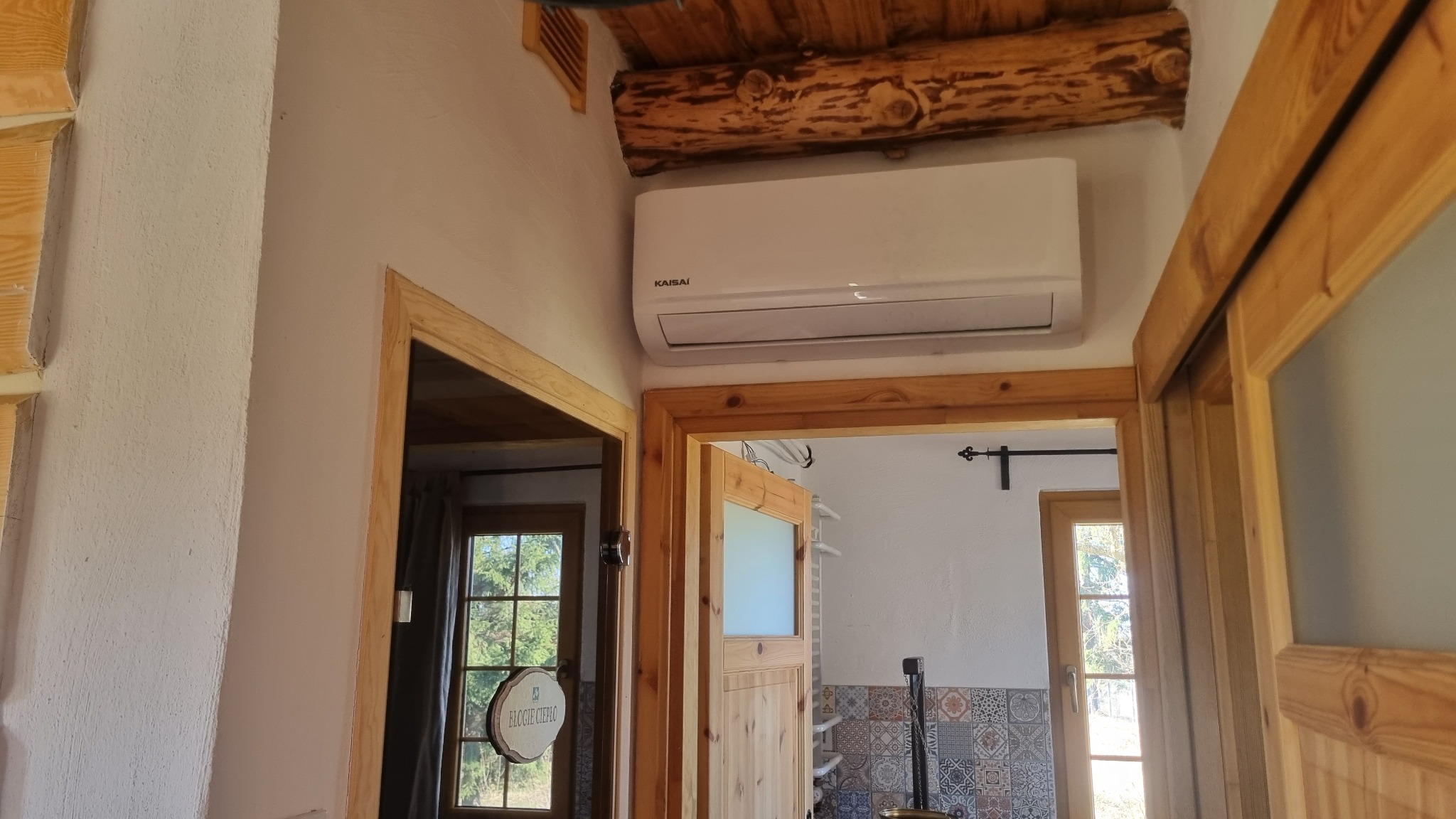 Biały klimatyzator marki Kaisai zamontowany na ścianie w drewnianym wnętrzu, widoczny nad framugą drzwi z naturalnego drewna.