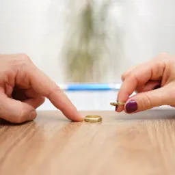 Dwie dłonie, męska i damska, zbliżają się do dwóch złotych obrączek leżących na drewnianym stole, w tle rozmyte dokumenty i roślina.