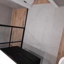 Nowoczesna łazienka z kabiną prysznicową typu walk-in z czarnymi profilami, umywalką na szafce w kolorze czarnym i płytkami imitującymi drewno na podłodze i ścianie.