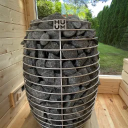 Wnętrze sauny z piecem na kamienie w metalowej obudowie, widok przez okno na zielony ogród.