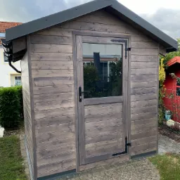 Drewniana, impregnowana szopa ogrodowa z oknem i czarnym dachem, widoczna na tle zieleni i kostki brukowej, z dekoracyjnymi zawiasami przy drzwiach.