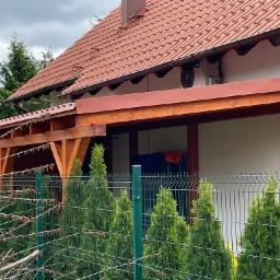 Zadaszenie tarasu z drewnianą konstrukcją, widoczne zza zielonego ogrodzenia i żywopłotu z tuj. Na ścianie budynku zamontowana klimatyzacja.