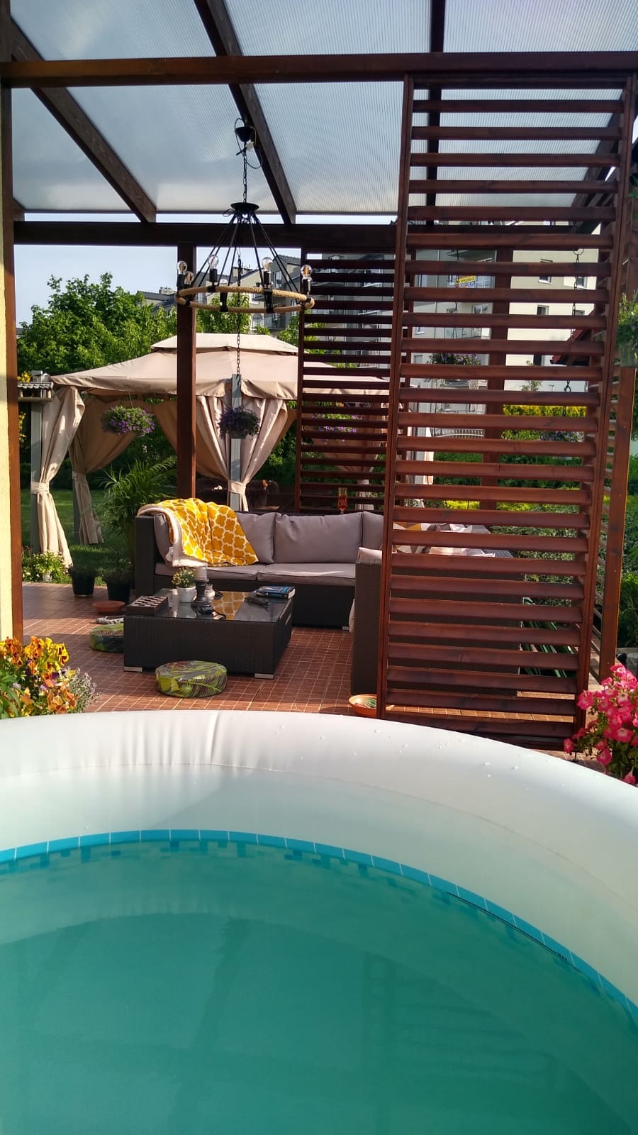 Zadaszona przestrzeń relaksacyjna z drewnianą konstrukcją, meblami ogrodowymi, wiszącą lampą i widokiem na basen.