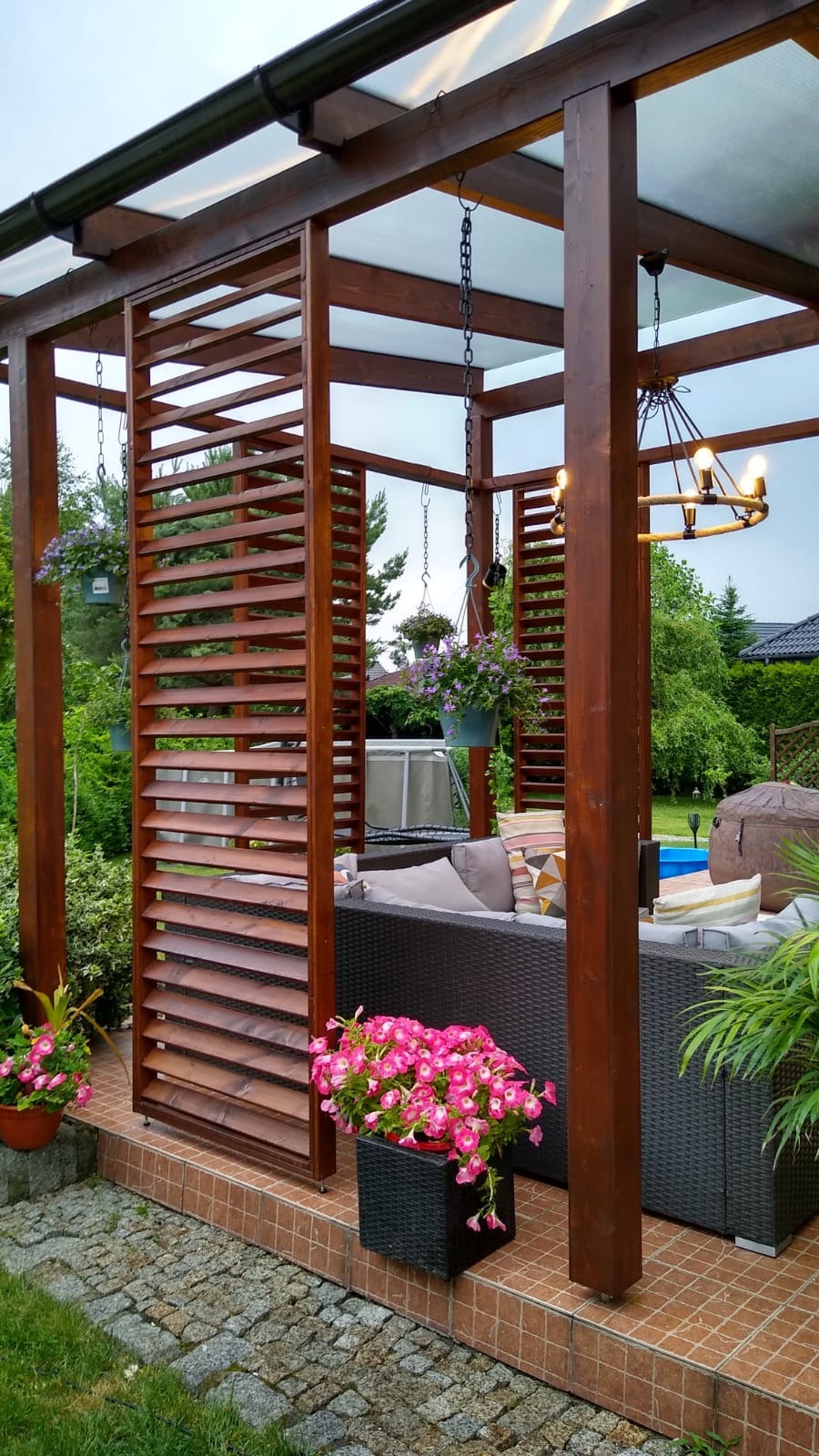 Zadaszona konstrukcja z drewnianymi lamelami, wyposażona w wiszące kwietniki i oświetlenie sznurowe, tworząca zaciszną strefę relaksu z rattanowym narożnikiem i dekoracyjnymi poduszkami, otoczona...