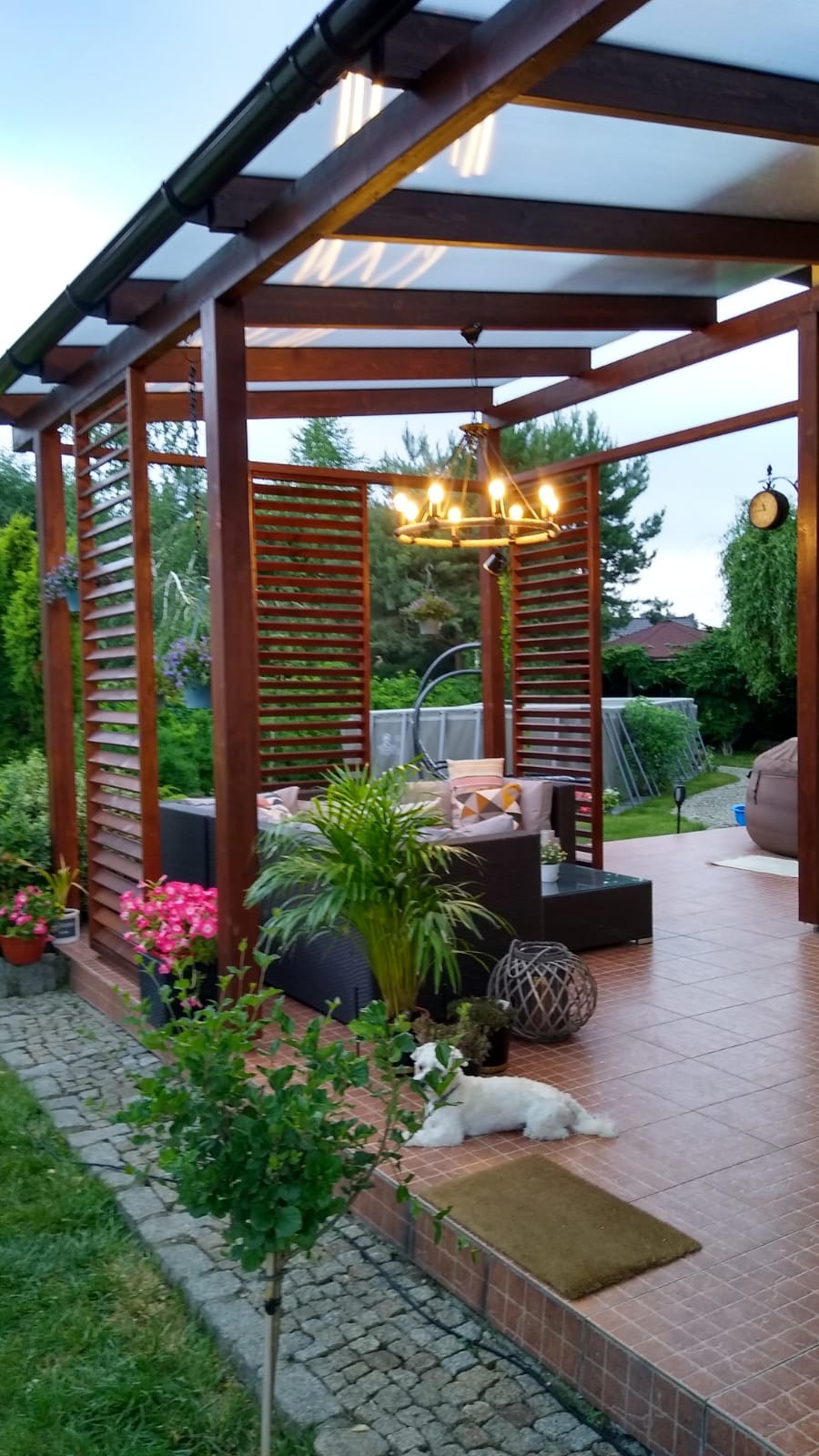 Zadaszona konstrukcja z drewnianymi panelami, oświetlona żyrandolem, z wypoczynkową sofą, roślinami doniczkowymi i białym psem leżącym na wycieraczce.